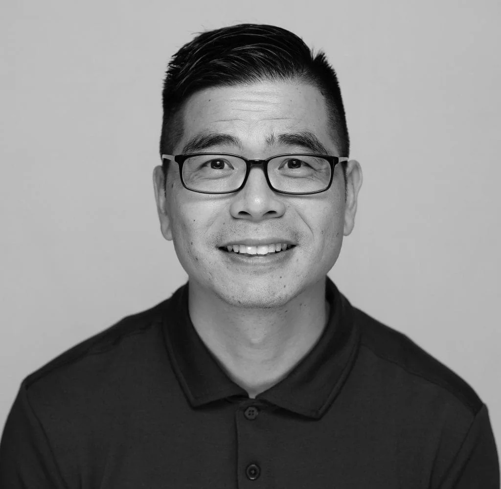 A black and white headshot depicting a man with close cropped dark hair, thin dark glasses, and a black shirt from the chest up against a plain gray background