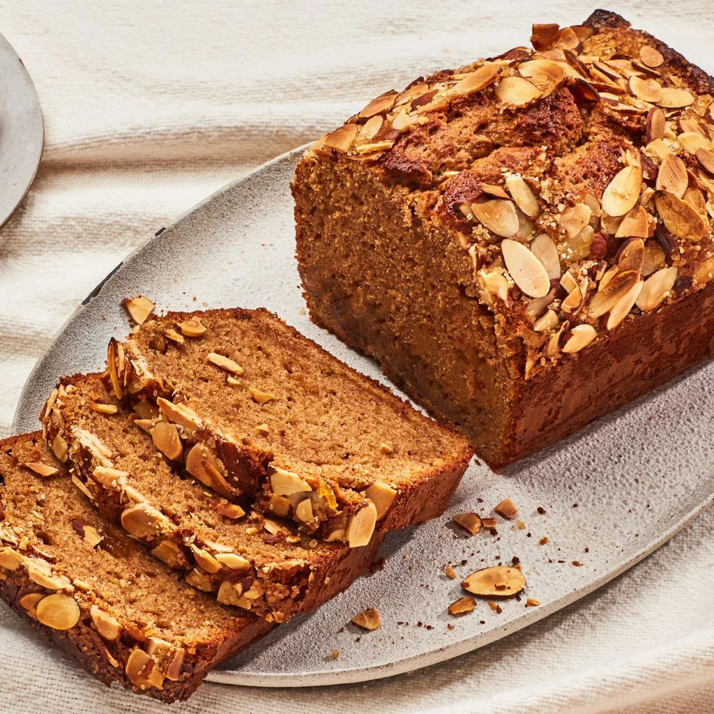 A loaf of almond butter quick bread topped with sliced almonds with several slices arranged on a platter and a fork...