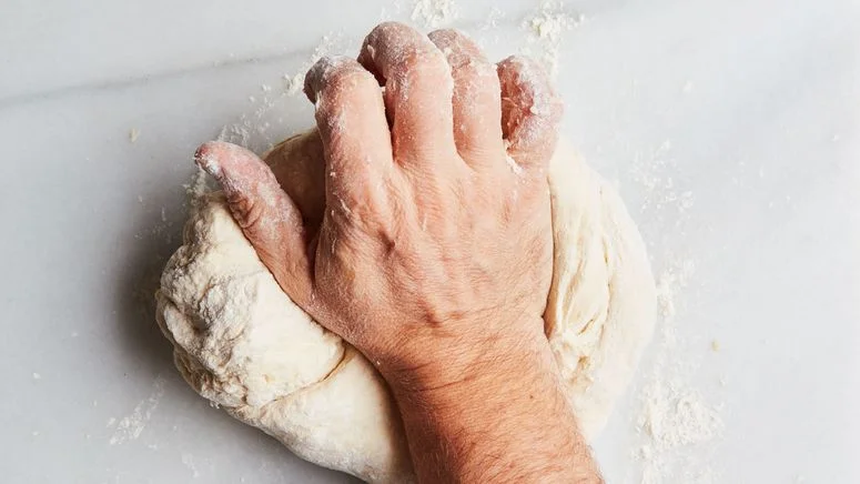A hand, kneading bread dough on a marble surface to demonstrate the difference in bread flour vs. all-purpose flour.