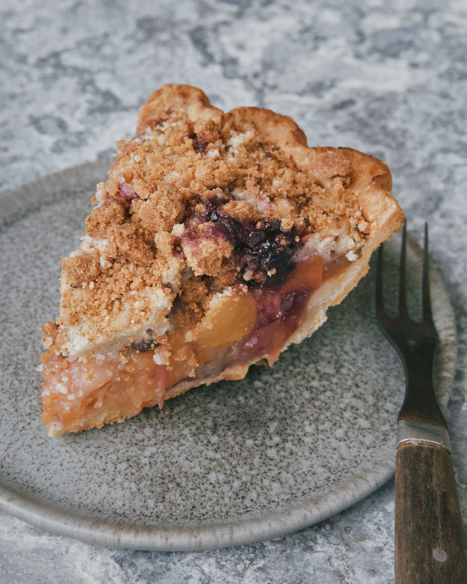 A slice of peach and blueberry pie with a cardamom crumble topping on a plate.