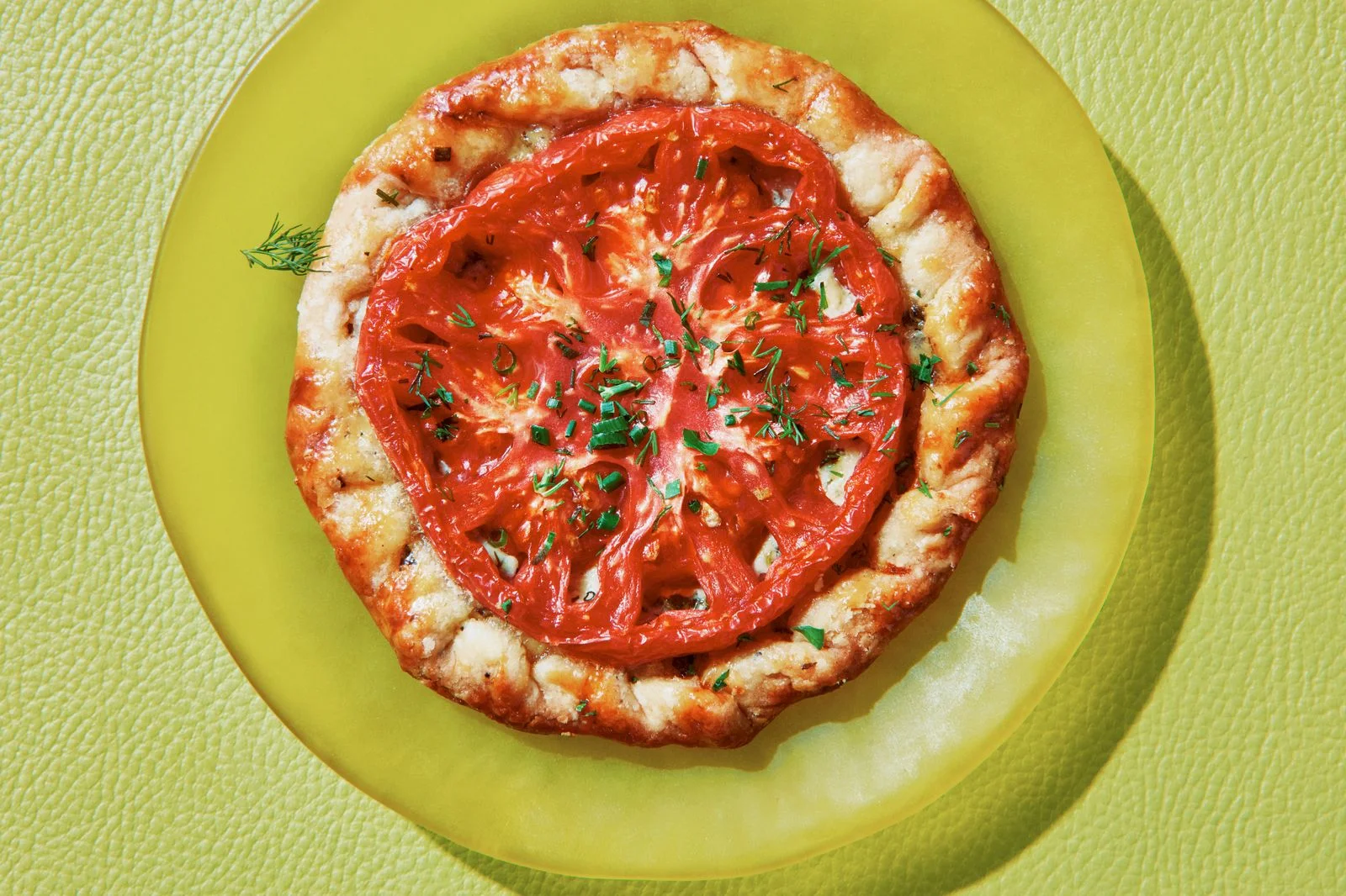 A round of flaky pastry topped with a sliced heirloom tomato chives and dill on a plate.