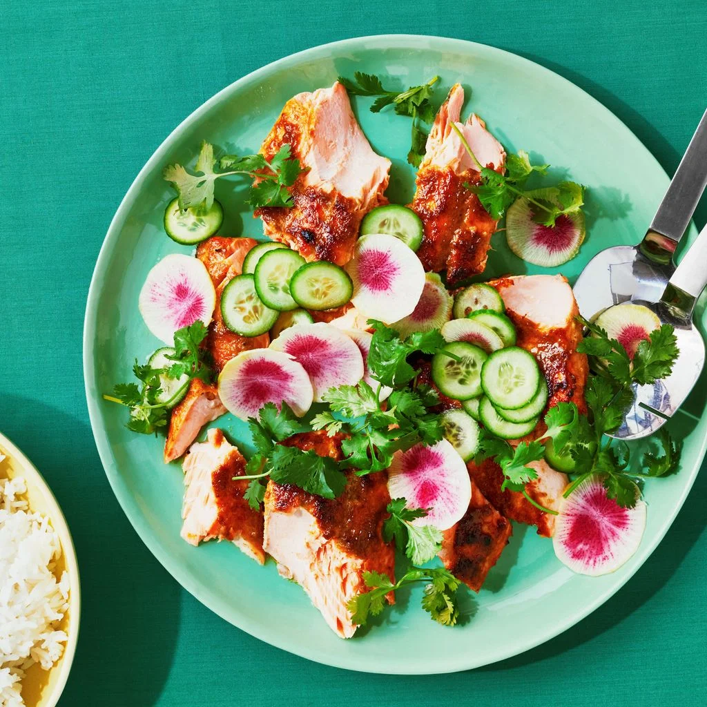 Broken up pieces of broiled salmon covered in a green curry sauce on a platter with sliced cucumbers rainbow radishes...