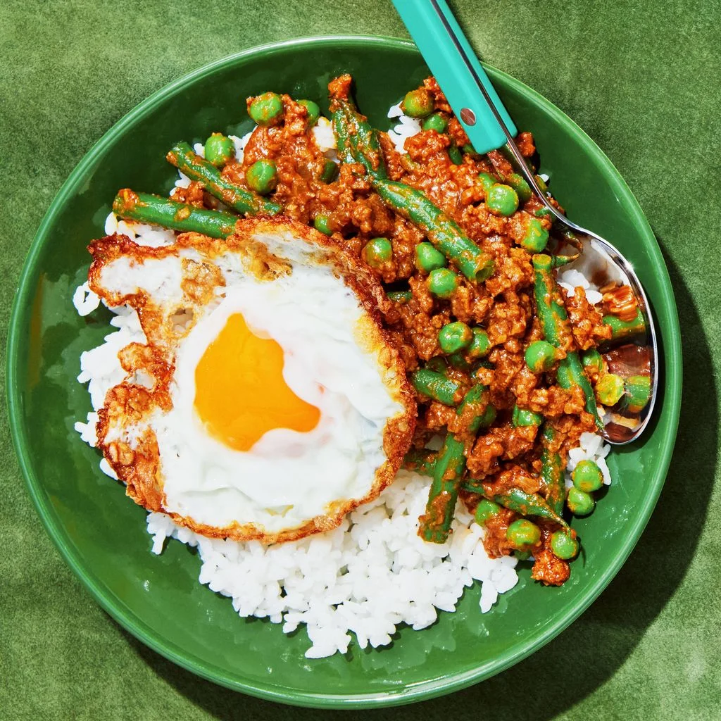 Ground beef cooked in curry with green beans and peas in bowls with white rice and fried eggs.