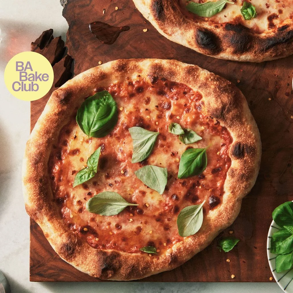 Two whole cheese pizze topped with red pepper flakes olive oil and basil leaves on a wooden cutting board.