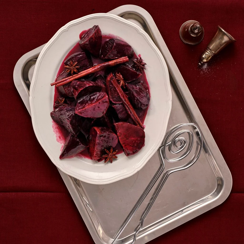 A white platter of saucy candied purple sweet potatoes interspersed with cinnamon sticks and star anise atop a silver...