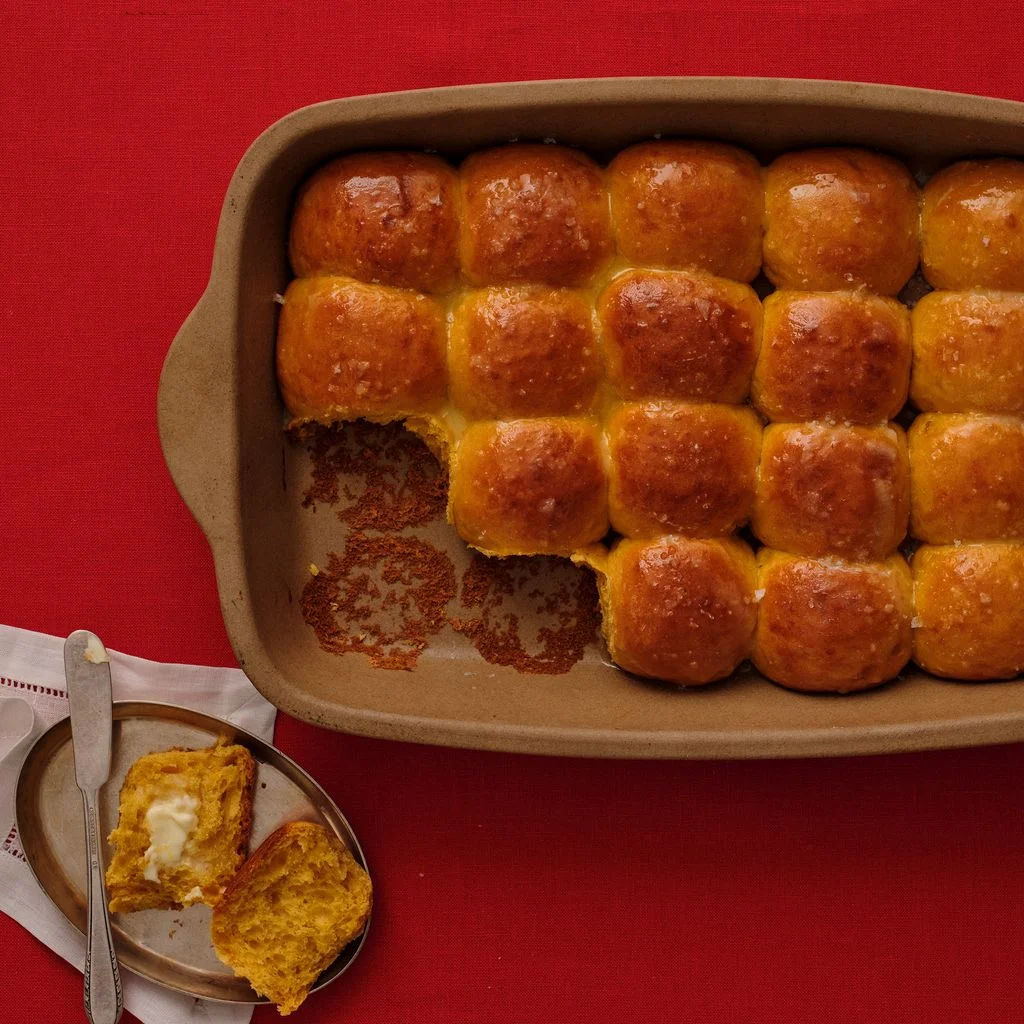 A neutralcolored stone baking dish of salted sweet potato rolls alongside a small dish with a halved roll buttered on...