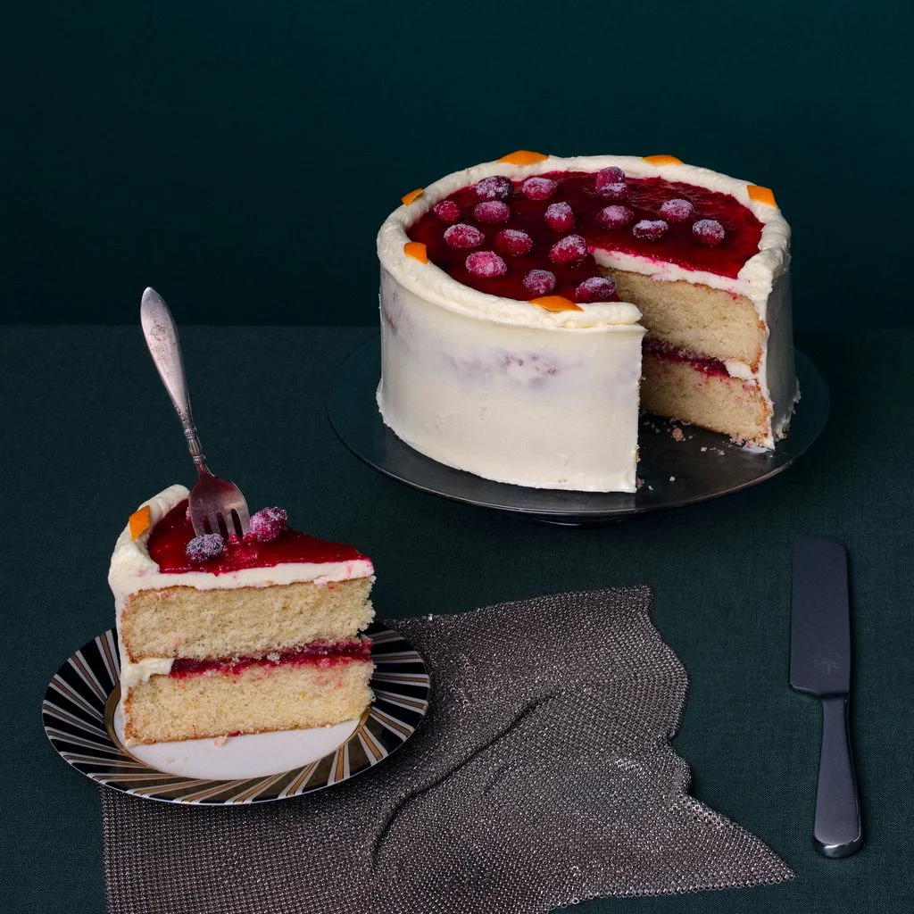A slice of a twolayer white chocolate frosted cake sits alongside the full cake topped with cranberry jam sugared...