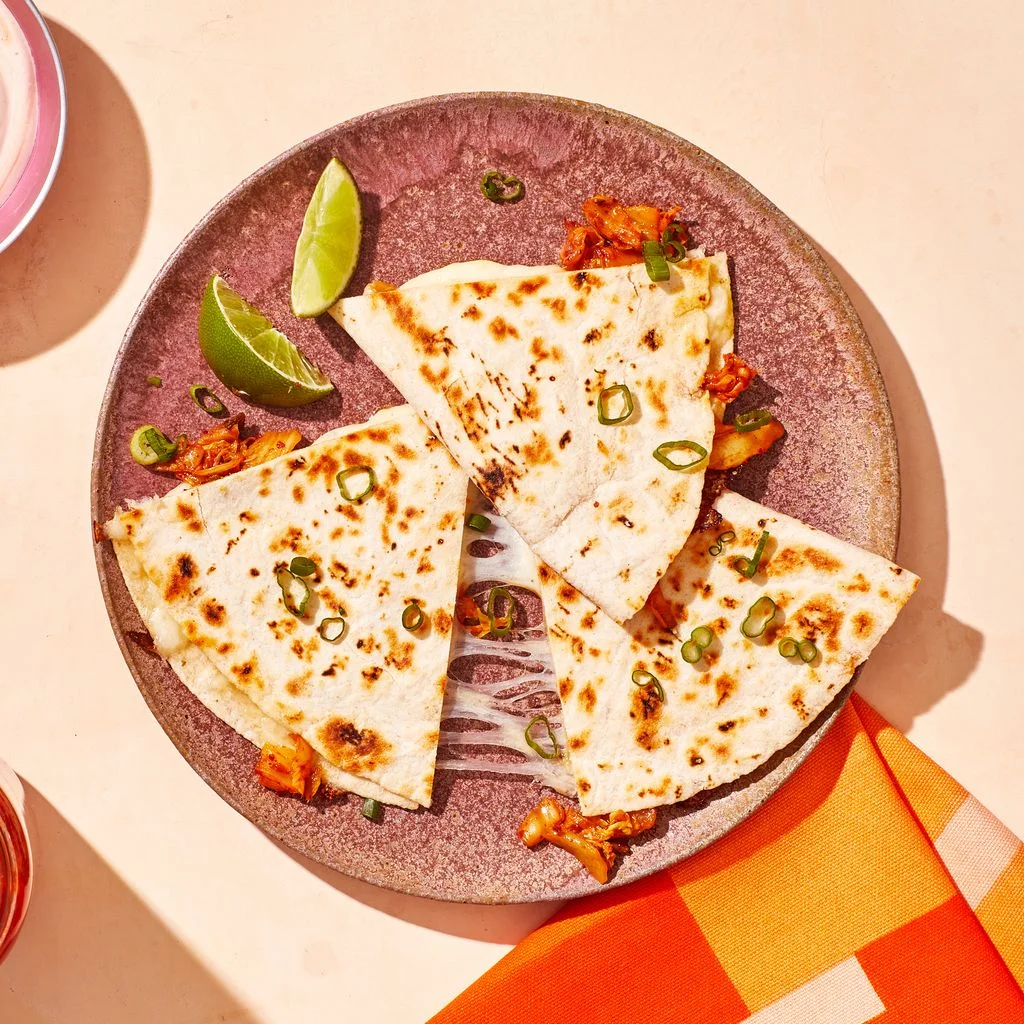 Three pieces of a kimchi quesadilla on a plate with lime wedges next to a jar of kimchi and a bowl of kimchisour cream...