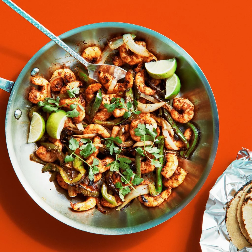 Sauted shrimp onions and green bell peppers in a skillet with cilantro and lime wedges with warm tortillas alongside.