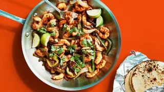 Sauted shrimp onions and green bell peppers in a skillet with cilantro and lime wedges with warm tortillas alongside.