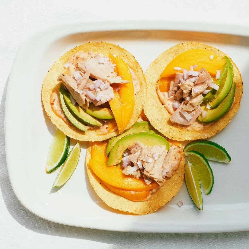 Three tostadas with canned tuna sliced mangoes sliced avocados shallots and spicy mayo on a platter served with lime wedges.
