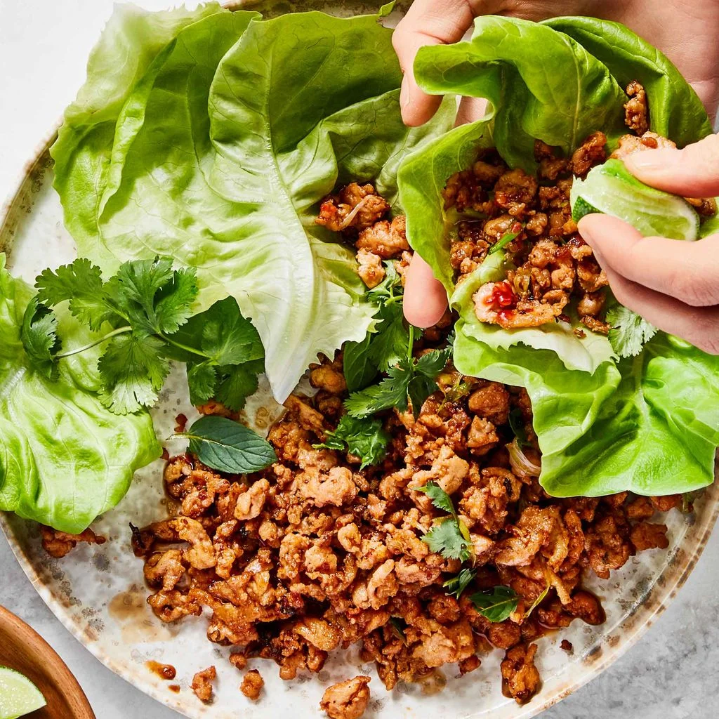 Two hands assembling a spicy chicken lettuce wrap with ground chicken cilantro mint and lime wedges.