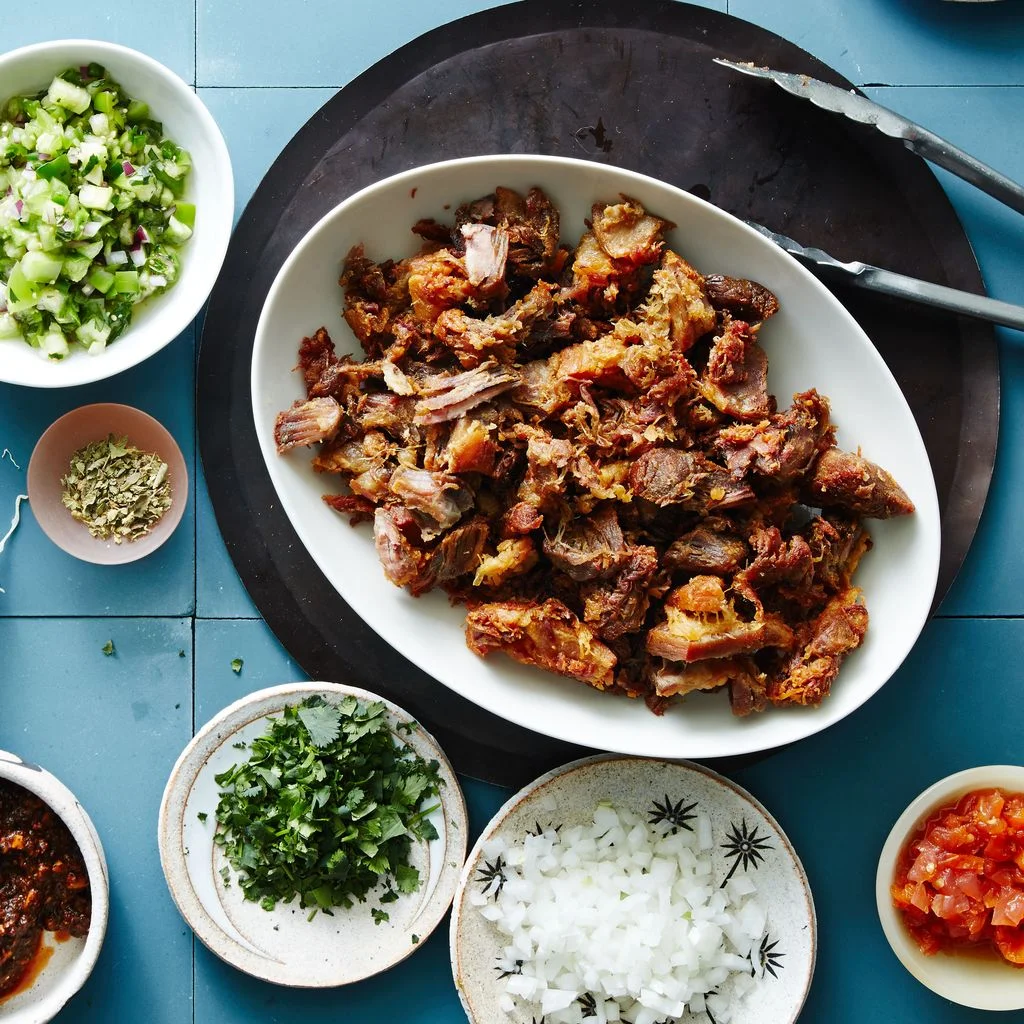 A big platter of pork carnitas surrounded by taco fixings including tortillas shredded cabbage salsa verde and more.