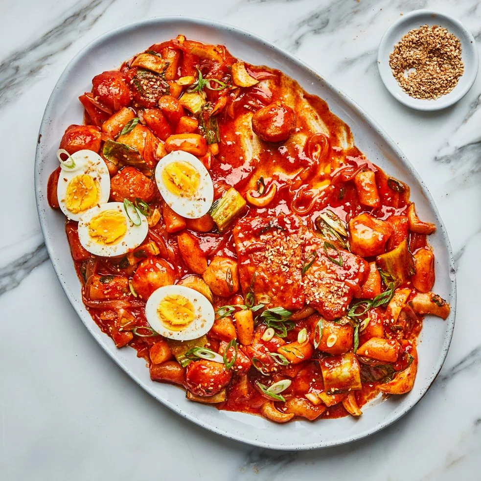 Tteokbokki a dish of Korean rice cakes enrobed in a spicy red gochujang sauce topped with halved boiled eggs on a platter.