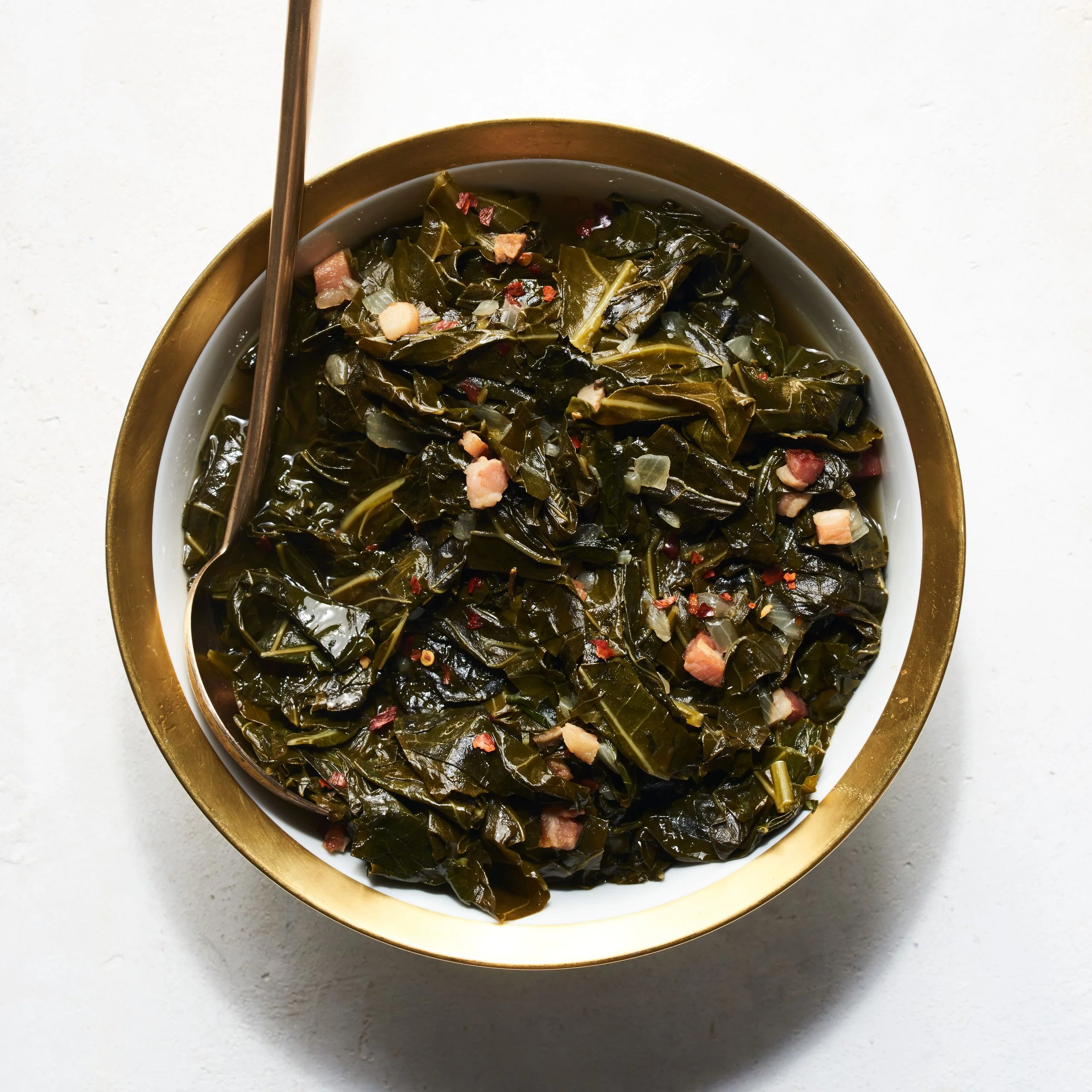 Braised collard greens with bacon in a serving bowl with a gold edge and gold spoon.