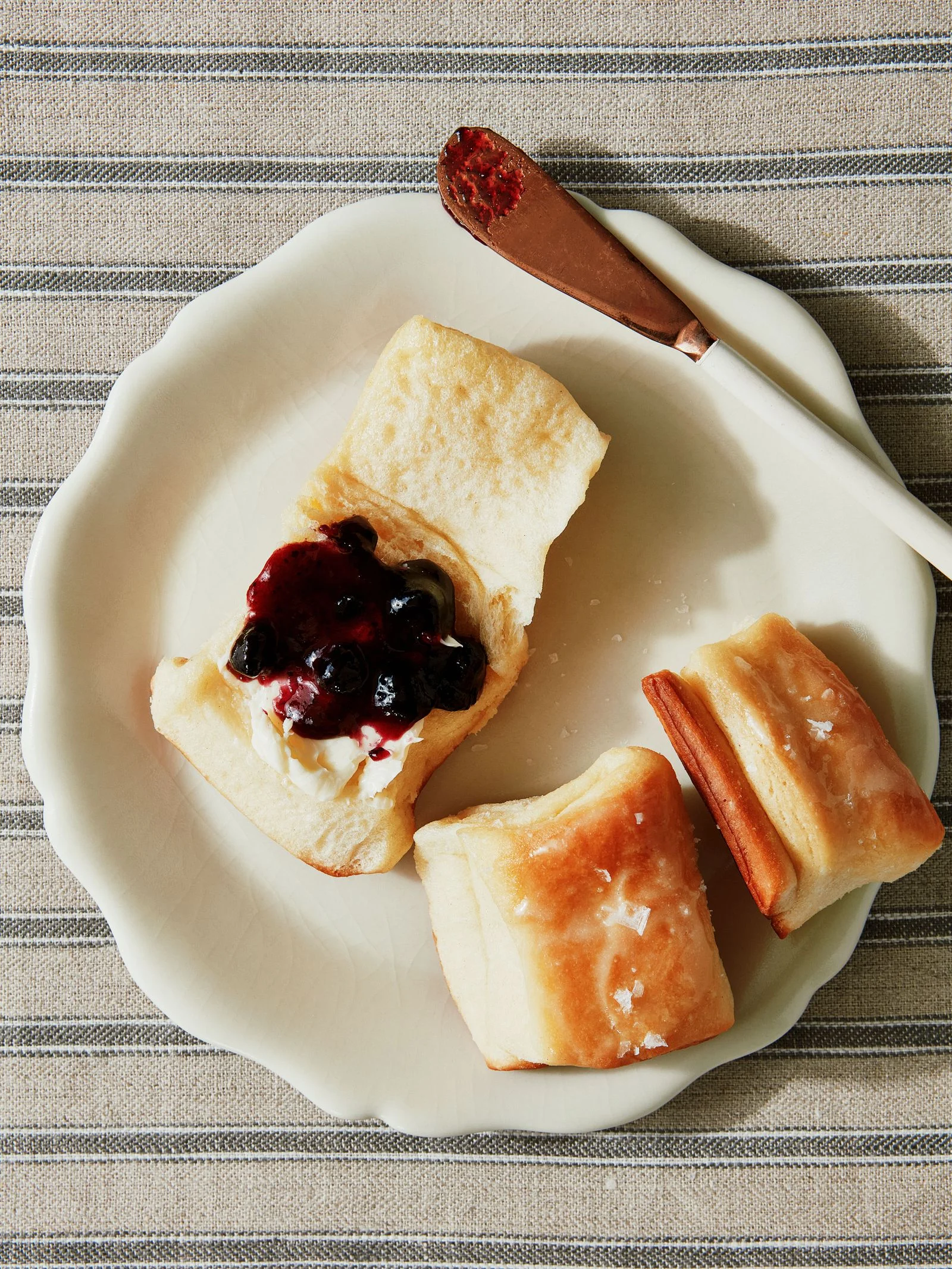 Three Parker House Rolls on a plate one opened with warm butter and blueberry jam on top.