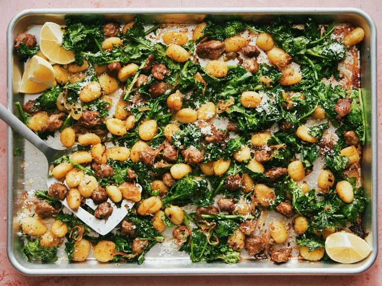 Gnocchi on a sheet tray with pieces of Italian sausage broccoli rabe red onions parmigiana and lemon wedges.
