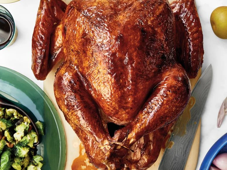 A glistening drybrined turkey resting on a cutting board surrounded by various Thanksgiving side dishes.