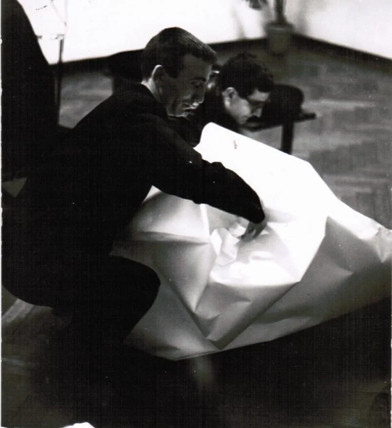 Black and white photograph of Agostino Bonalumi crouching on the floor and working on a large white shaped canvas at Galleria Eidac, 1962, with a second figure visible in the background. Courtesy Archivio Bonalumi.