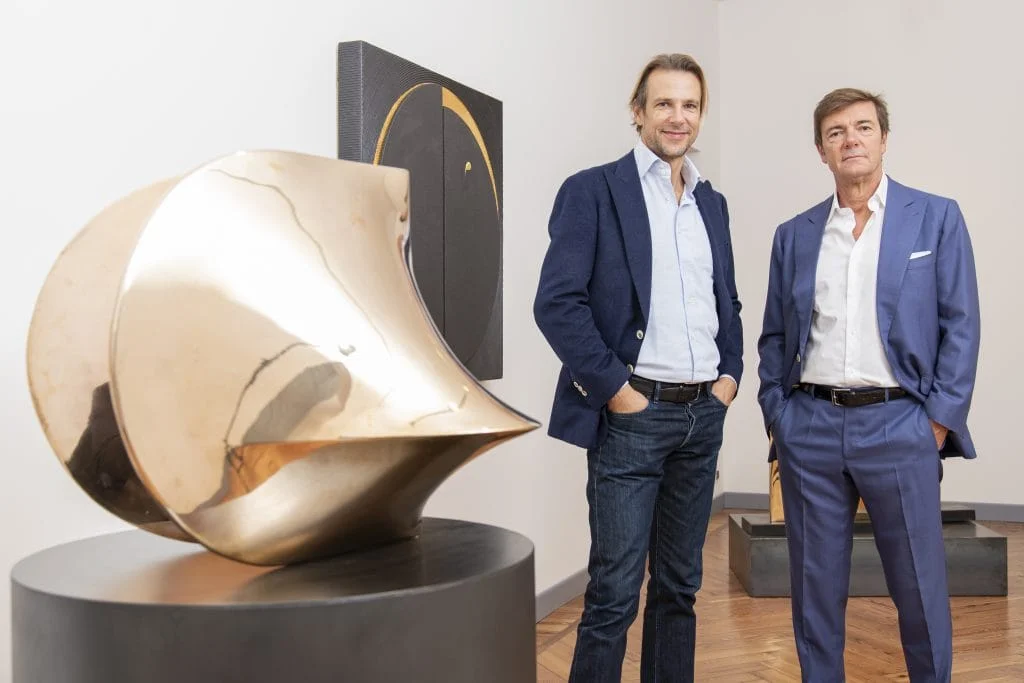 Two men standing in an art gallery beside a large polished bronze abstract sculpture on a dark pedestal, with a dark geometric painting hanging on the wall behind them. The gallery features warm wood parquet flooring.