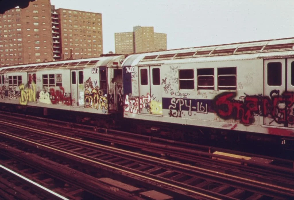 Heavily graffiti-covered New York City subway cars on an elevated track in the 1970s or 80s, with residential apartment buildings visible in the background