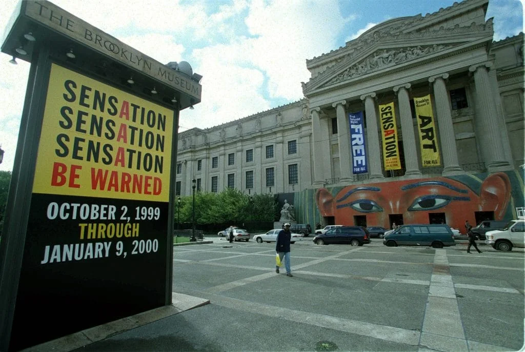 Brooklyn Museum exterior displaying signage and banners for the controversial 'Sensation' exhibition, running October 2, 1999 through January 9, 2000, with a large painted face mural on the facade