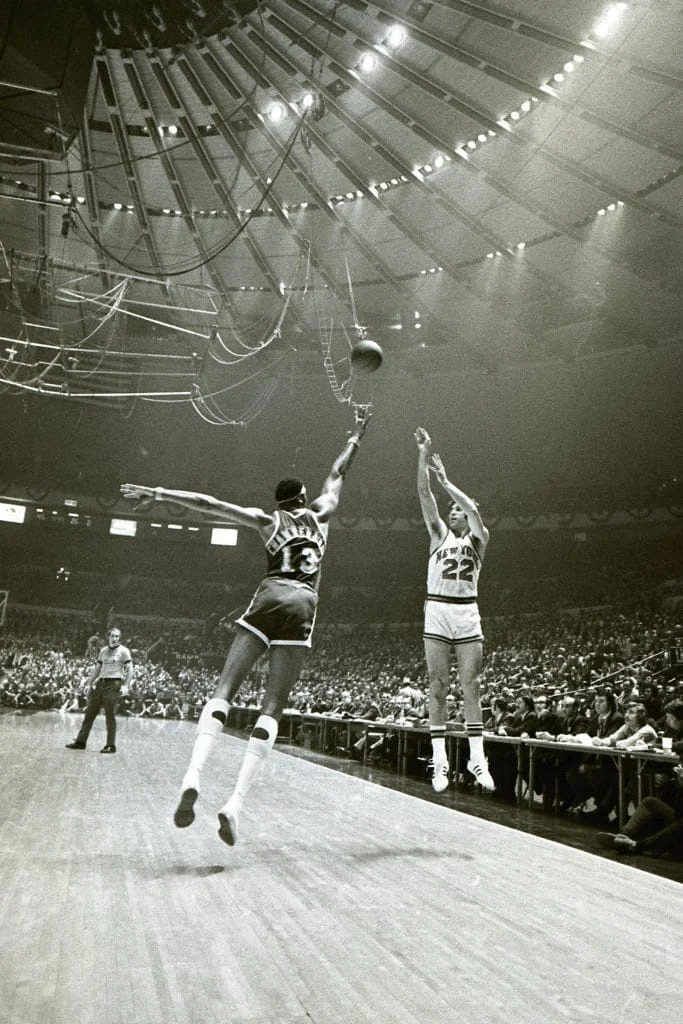 Black and white photo of New York Knicks number 22 shooting over a defender number 13 in a packed 1970s NBA arena