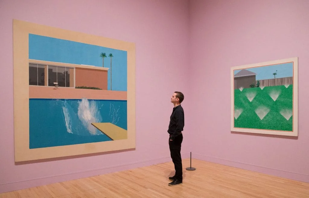 A photograph of a man in black pants and black shirt standing in a pink-walled museum gallery looking at an iconic David Hockney painting of a Hollywood pool with a splash in it, while another painting of a green lawn punctuated by sprinklers hangs on the adjoining wall, behind him