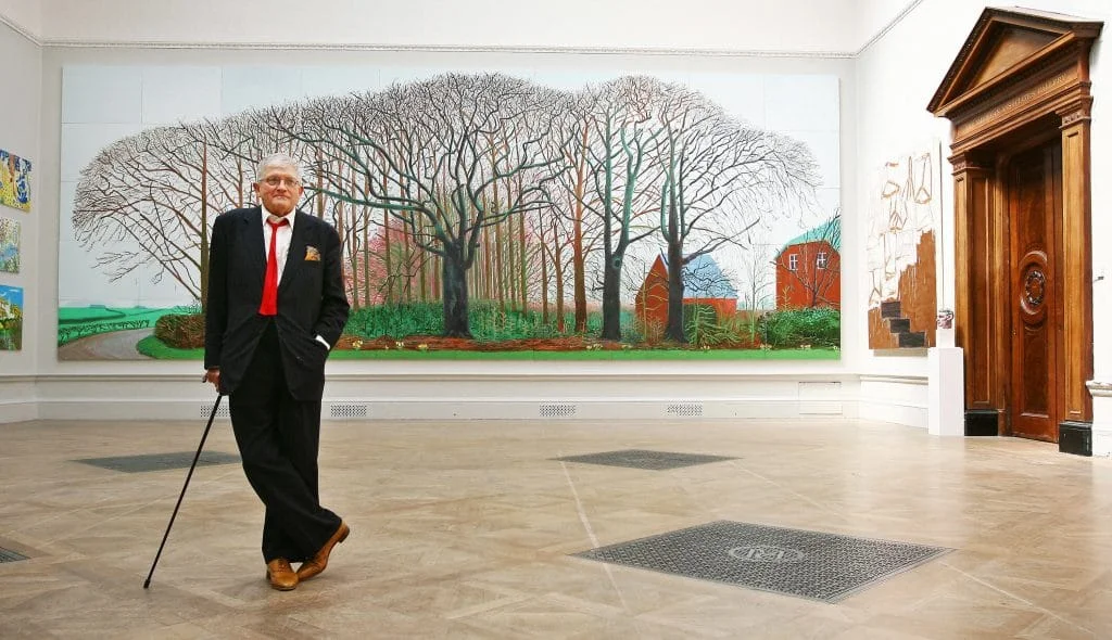 A photograph of David Hockney taken nearly two decades before his death, standing in a black suit with white shirt, red tie, and a cane before a massive painting featuring a landscape of tall, barren trees
