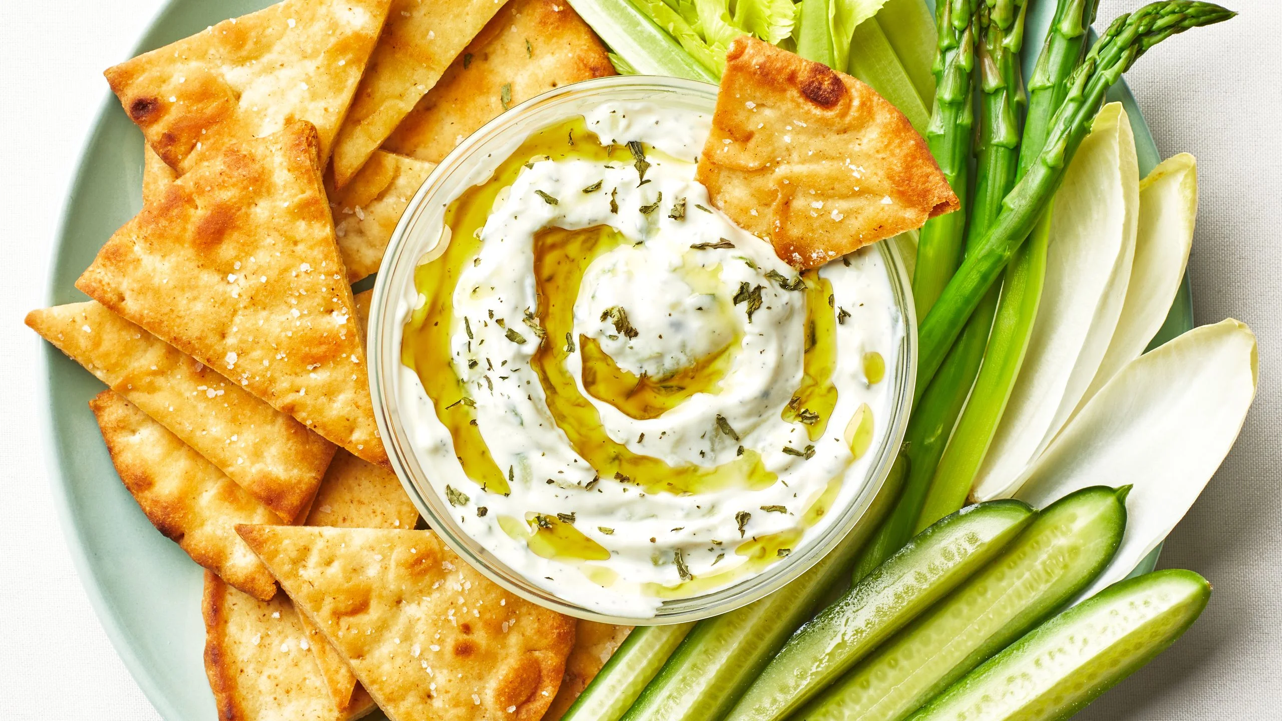 Bowl of homemade tzatziki drizzled with olive oil and surrounded by pita chips cucumber spears endive and asparagus.