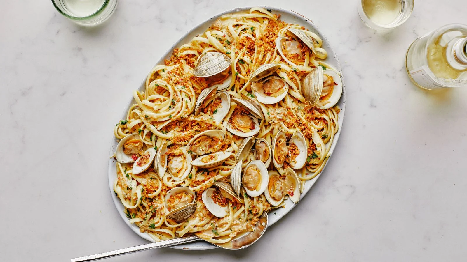 A large platter of linguine with clams alongside a bottle of white wine and two glasses.