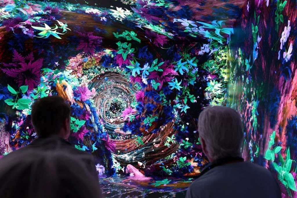 Two people observe an immersive, swirling digital projection of vibrant, glowing flowers and dark wood textures creating a deep, tunnel-like effect