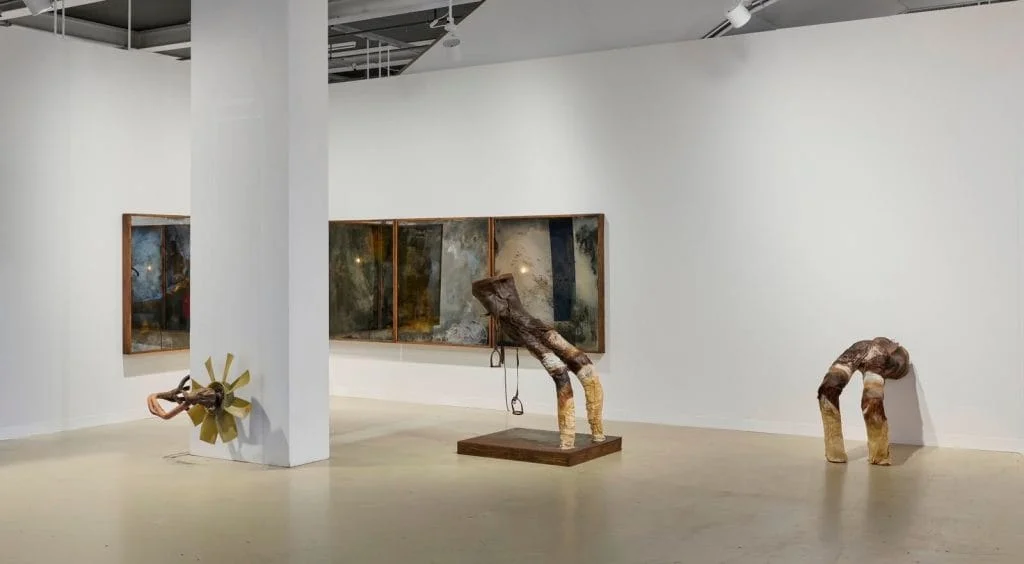 a white walled space at Art Basel contains artworks, some hang on the wall and others are sculptures, they are brown and yellow toned