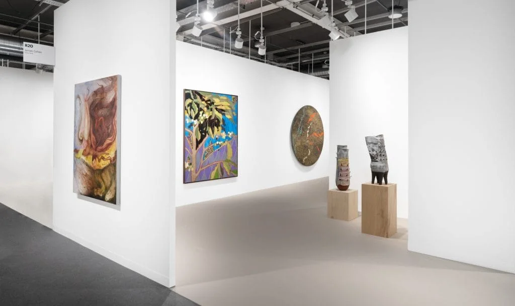 a white walled booth space at Art Basel is filled with artworks on the walls and on wooden podium