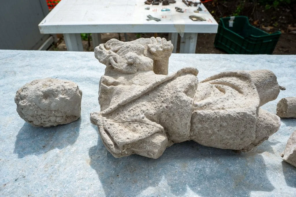 A photograph of a fragmented white statuette of a man holding an animal laying in pieces on a table, with another table in view behind it