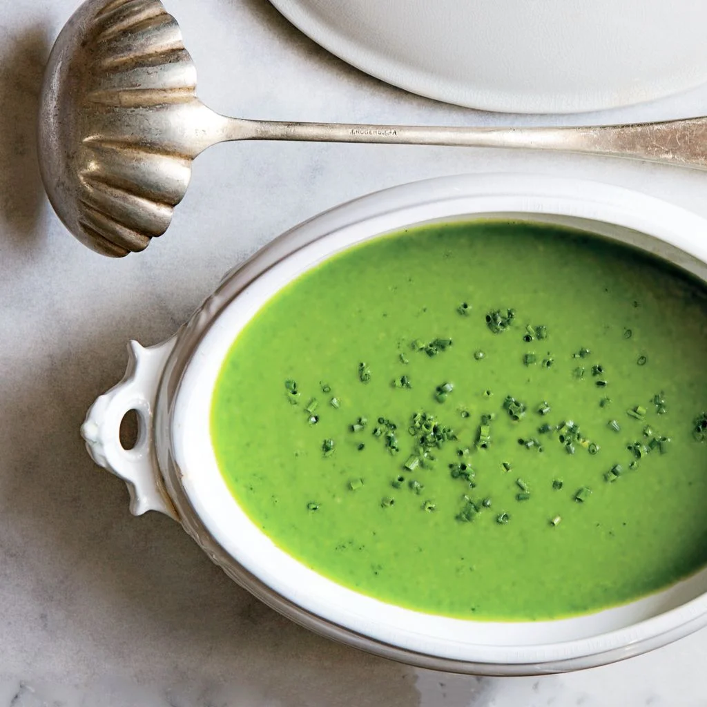 A tureen of bright green mintandgreenpea soup garnished with chives with a ladle for serving.