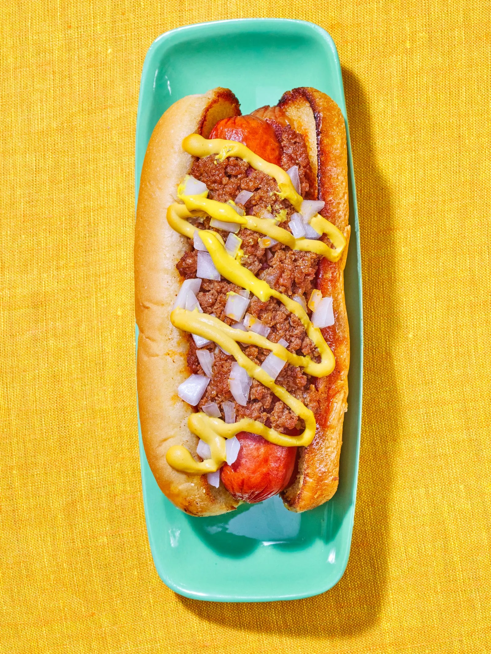 A hot dog loaded with chili onions and mustard on top of a slim green plate.