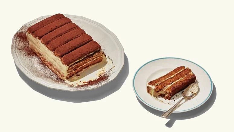 A loaf of tiramisu topped with cocoa dusted ladyfingers with one serving removed to a plate.