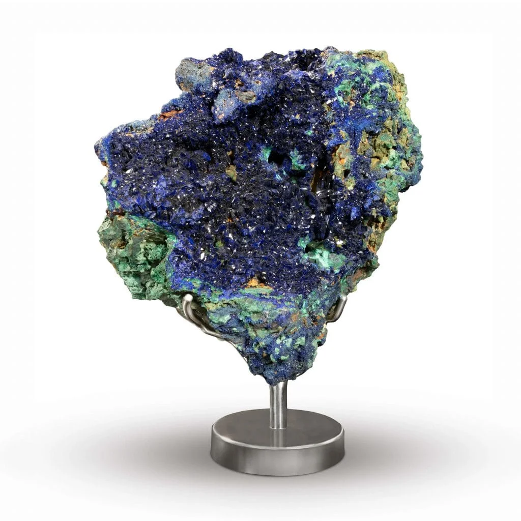 A photograph of a sturdy, textured hunk of iridescent minerals in shades of blue with a few specks of yellow sitting atop a metal stand before an all-white background