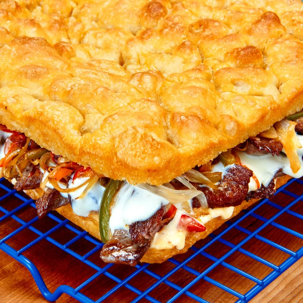 A largeformat focaccia sliced in half and stuffed with cheesesteak fixings all atop a blue wire rack.