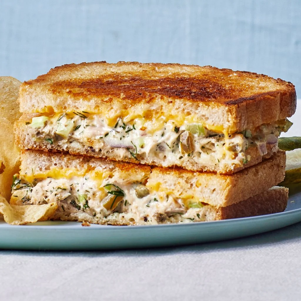 Two halves of a classic tuna melt sandwich stacked on a plate with potato chips covered in Frank's red hot and a pickle...