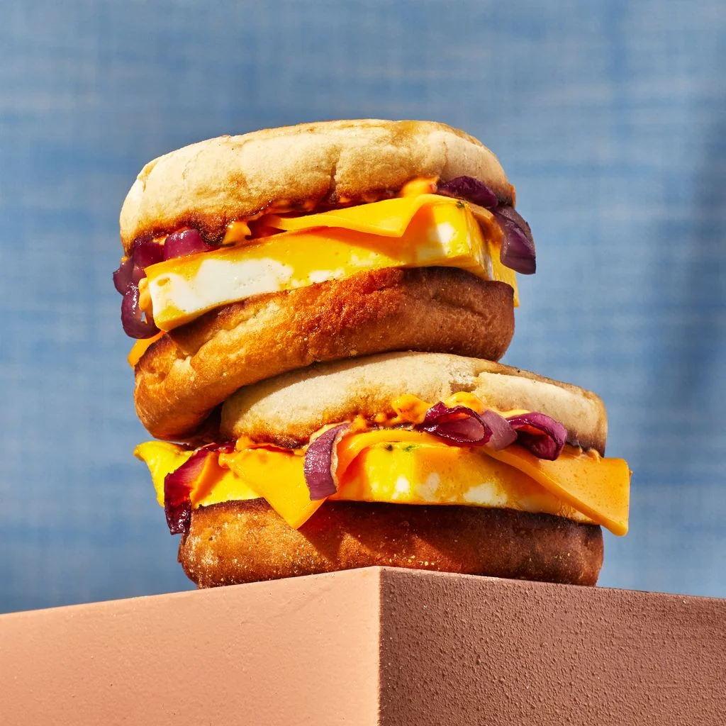 Two egg and cheese breakfast sandwiches on English muffins stacked on a wooden table.