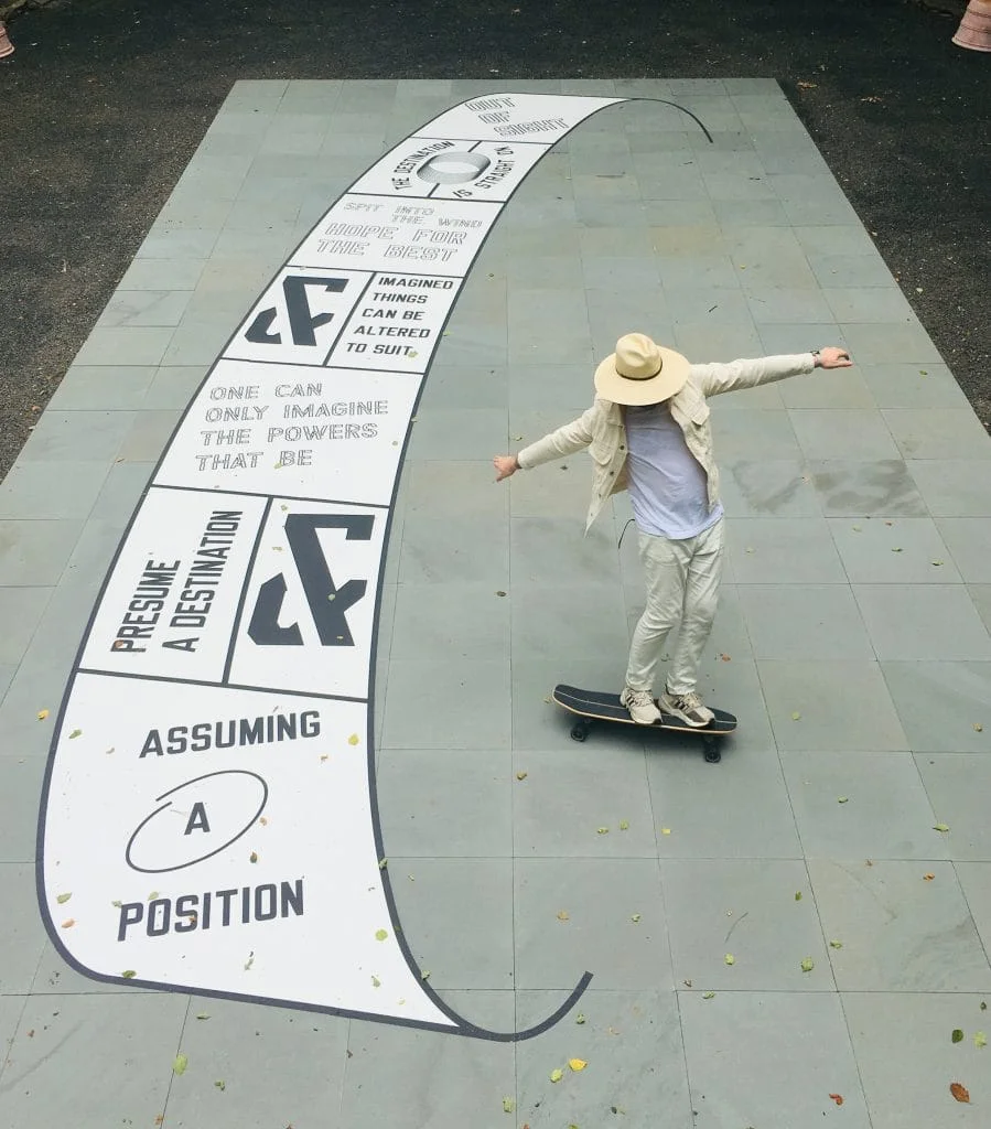 An aerial photograph of a man in a tan hat skateboarding with his arms out on a slab of pavement featuring a text-based black and white artwork composed in the shape of an unfuled scroll painted on the ground